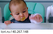 A hungry baby taps his hand on the table on a high chair, asks to eat, nibbles on a spoon. Teething, whims, itchy gums, introduction of complementary foods. Стоковое видео, видеограф Ольга Симонова / Фотобанк Лори