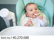 A hungry baby is gnawing on a plastic spoon at the table on a high chair. Teething, whims, itchy gums, introduction of complementary foods. Стоковое фото, фотограф Ольга Симонова / Фотобанк Лори