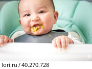 A dirty, joyful baby at the table on a feeding chair got his mouth dirty in vegetable puree. Introduction of complementary foods, the child likes to eat. Portrait close-up. Стоковое фото, фотограф Ольга Симонова / Фотобанк Лори