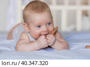 A charming boy in a bright bedroom. A newborn baby is lying on the bed and gnawing on toys. Teeth are teething. The baby's gums are itching. A child of 4 months looks into the camera. Family, pregnancy and childbirth. Стоковое фото, фотограф Photojuli / Фотобанк Лори