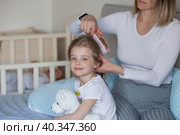 A happy young mother combs her adorable little daughter's hair while sitting in bed in the morning. The girl smiles. Family and care. Стоковое фото, фотограф Photojuli / Фотобанк Лори