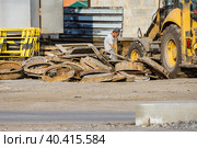 Kemerovo, Russia - june 2020 old manholes of wells are piled in heap, road equipment and worker in cap sits in profile. Редакционное фото, фотограф Дмитрий Коростылев / Фотобанк Лори