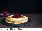 Pile of russian homemade traditional blinis, also crepes with red berries served on round stone tray ready for eating on dark brown wooden table against black background. Image with copy space. Стоковое фото, фотограф Елена Кабенкина / Фотобанк Лори