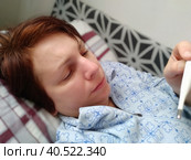 Sick woman in bed. Headache, runny nose and cold. Paleness of the face and faintness of the body. Feeling unwell. The patient lies on a pillow, holds a thermometer and measures the temperature. Стоковое фото, фотограф Юлия Петровская / Фотобанк Лори