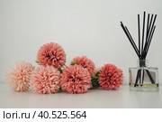 Composition with incense sticks, essential oil in a glass vase and pink terry artificial flowers in the interior of a white room. White background. Lots of flowers and scent sticks. Стоковое фото, фотограф Юлия Петровская / Фотобанк Лори