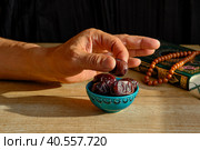 Hand of senior Muslim man taking dates from bowl on background of book of Quran and rosary. Стоковое фото, фотограф Ирина Кононова / Фотобанк Лори