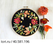 A bright floral ornament of orange, red and gold flowers is embroidered with satin ribbons on black velvet and a bouquet of autumn flowers next to it on a light wooden table. Flat lay, close-up. Стоковое фото, фотограф Виктория Катьянова / Фотобанк Лори