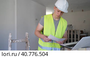 Fifteen year old worker in a yellow vest checks the house repair plan on a laptop, holding papers with drawings in his hands. Стоковое видео, видеограф Яков Филимонов / Фотобанк Лори