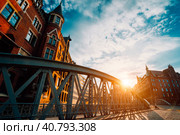 Metal arch bridge and old red bricks building in the Speicherstadt warehouse district of Hamburg HafenCity with sunburst light during sunset golden hour and white clouds against blue sky above (2018 год). Стоковое фото, фотограф Igor Tichonow / Фотобанк Лори