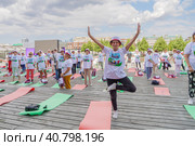 Женщина пенсионерка в фирменной футболке и шапочке стоит в йоговской асане на фоне других пенсионеров, готовящихся к спортивным занятиям в парке Музеон на Крымской набережной (2023 год). Редакционное фото, фотограф Сергей Никифоров-Гордеев / Фотобанк Лори
