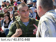 Десантник в тельняшке, в полевой одежде и с автоматом за спиной выслушивает наставления от ветерана ВДВ. Действие происходит 2 августа во время празднования дня ВДВ. Сзади видны зрители на стадионе. На некоторых одеты голубые береты. Редакционное фото, фотограф Сергей Никифоров-Гордеев / Фотобанк Лори