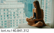 Young smiling woman drinking delicious coffee near window with big city skyscrapers outside. Relaxed girl enjoys morning cappuccino on weekend in the metropolis. Slow motion close-up footage. Стоковое фото, фотограф Ольга Сухарева / Фотобанк Лори