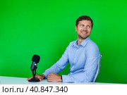 Portrait of handsome man broadcaster with microphone at desk on green backdrop, looking away. Smiling male broadcaster in television studio, lifestyle. Tele production concept. Copy ad text space. Стоковое фото, фотограф Aleksandr Golubev / Фотобанк Лори