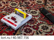 Бюджетная игровая приставка 8 Bit Junior с игровым картриджем лежит на советском ковре перед телевизором в городе Москве, Россия (2023 год). Редакционное фото, фотограф Николай Винокуров / Фотобанк Лори