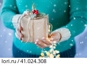A woman in a green sweater holds a craft paper box in her hands with a Christmas decor made from a fir branch and red berries. Close-up of female hands holding christmas or new year gift. Snowfall. Стоковое фото, фотограф Оксана Прахова / Фотобанк Лори
