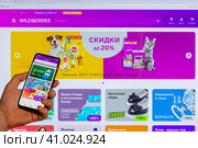 Adygea, Russia - August 09, 2023: hand man with smartphone making purchases in Wildberries online store in mobile application against background Wildberries marketplace web page open computer monitor. Редакционное фото, фотограф Андрей С / Фотобанк Лори
