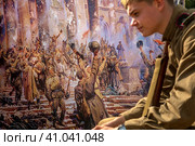 Молодой парень с винтовкой в форме времен Великой Отечественной войны сидит на фоне картины художник П. Кривоногова "Победа" (1948 год) в городе Москве, Россия (2023 год). Редакционное фото, фотограф Николай Винокуров / Фотобанк Лори