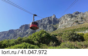 The funicular ascends Table Mountain in Cape Town. Against the background of the blue sky, cables, a red round booth. The cliff slopes are steep and rocky. Green vegetation on the ground. South Africa. Стоковое фото, фотограф Вера Ивановна Кубарева / Фотобанк Лори