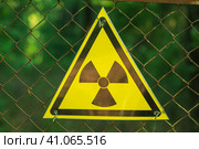 Close-up of a yellow triangular radiation sign on a wire mesh. In the background is a green forest. Warning of danger. Стоковое фото, фотограф Воловодов Владимир / Фотобанк Лори