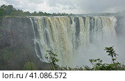 The magnificent Victoria Falls. The Zambezi River flows from the edge of the plateau into the gorge. Fog over powerful streams of water. There is green vegetation in the foreground. Zimbabwe (2020 год). Стоковое фото, фотограф Вера Ивановна Кубарева / Фотобанк Лори