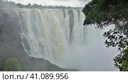 The Zambezi River flows from the edge of the plateau into the gorge. There is a thick fog over the abyss. In the foreground are green tree branches. Victoria Falls. Zimbabwe. (2020 год). Стоковое фото, фотограф Вера Ивановна Кубарева / Фотобанк Лори
