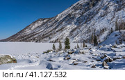 The surface of the frozen lake is covered with pure untouched snow. Picturesque boulders are scattered on the shore. A chain of footprints trampled in snowdrifts. Mountain slope against the blue sky. Стоковое фото, фотограф Вера Ивановна Кубарева / Фотобанк Лори