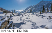 Stones under a layer of snow are scattered across the valley. The chain of footprints is trampled in snowdrifts. In the distance -three people walking forward. Coniferous trees, mountains. Altai. Стоковое фото, фотограф Вера Ивановна Кубарева / Фотобанк Лори