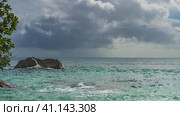 A granite boulder rises above the surface of the turquoise ocean. Foam waves on the rocks. Rain clouds in the sky. Green tree branches in the foreground. Seychelles. Стоковое фото, фотограф Вера Ивановна Кубарева / Фотобанк Лори