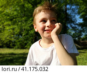 A boy looks into the distance, thinks and smiles. The child propped his chin in his hand and frowned. The process of thinking and a half smile. In the background is green grass and trees. Стоковое фото, фотограф Юлия Петровская / Фотобанк Лори