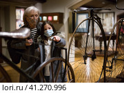 Tween schoolgirl and elderly female tutor in face masks viewing vintage bicycle in museum. Стоковое фото, фотограф Яков Филимонов / Фотобанк Лори
