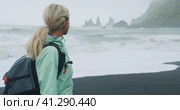 Woman with a green raincoat walking on a black sand beach enjoying view to Reynisfjara sea stack cli. Стоковое фото, фотограф Igor Tichonow / Фотобанк Лори