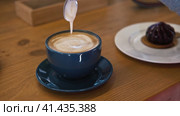 A Hand is Mixing the Milk Foam of Latte. Person Stirring Cappuccino with Steel Spoon. Hot Coffee in a Dark Blue Ceramic Cup on a Wooden Table. Modern Coffee shop, Cafeteria. Thick Froth. Close-up View. Стоковое видео, видеограф Марина Демидюк / Фотобанк Лори