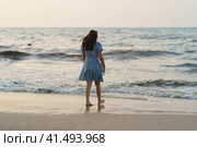 Zelenogradsk, Russia - August 24, 2022: Photography of the amazing Baltic sea. Waves going onto sandy beach. Beautiful young woman standing on the beach. The wind blows her hair and light dress. Concept of beauty in nature. Back, rear view. Редакционное фото, фотограф YuryKara / Фотобанк Лори