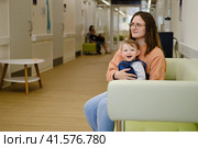 A mother woman with a toddler baby on a couch in the corridor of the clinic. Mom and child in the lobby of the polyclinic. Kid aged one year. Стоковое фото, фотограф Андрей Журавлев / Фотобанк Лори