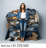 A young attractive girl of about 20 years old is standing in a blouse and jeans in front of a large pile of clothes on a white background. Generated by AI. Стоковое AI-изображение, AI-художник Ярославский Максим / Фотобанк Лори