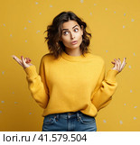 A young, pretty, attractive and thoughtful European girl of 20 years old in a gray casual T-shirt, arms spread apart, isolated on a uniform yellow background. Created by AI. Стоковое AI-изображение, AI-художник Ярославский Максим / Фотобанк Лори