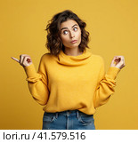 A young, pensive, insecure, rather attractive European girl of 20 years in a yellow casual blouse, spreads her hands questioningly, looking away. Created by AI. Стоковое AI-изображение, AI-художник Ярославский Максим / Фотобанк Лори