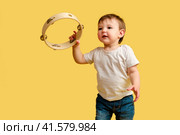 Toddler baby plays the tambourine, a child with a percussion musical instrument on a studio yellow background. Happy child musician playing hand drum. Kid aged one year four months. Стоковое фото, фотограф Андрей Журавлев / Фотобанк Лори