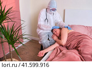 Doctor in protective medical uniform comforts and regrets sick adult woman on home bed, Стоковое фото, фотограф Андрей Журавлев / Фотобанк Лори