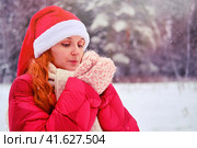 A woman with red hair blows on her frozen hands in knitted mittens. Redhead woman on Christmas and New Year Eve walks outdoors in a red hat and jacket. Стоковое фото, фотограф Андрей Журавлев / Фотобанк Лори