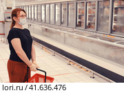 Adult woman in a medical mask stands in a department with frozen foods in a hypermarket. Sale of convenience foods and chilled food in the store. Flu virus quarantine in public places. Стоковое фото, фотограф Андрей Журавлев / Фотобанк Лори