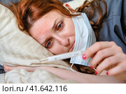 The girl with red eyes, sick with the flu, looks sadly at the thermometer. Woman lies on a bed in a medical mask. Стоковое фото, фотограф Андрей Журавлев / Фотобанк Лори