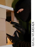 A man in a closed hood steals a box with a parcel in a warehouse at night. Concept of problems with theft of goods and postal parcels. Стоковое фото, фотограф Андрей Журавлев / Фотобанк Лори