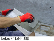 A male builder applies adhesive tile to lay it on the enclosing wall of the building from the outside. A construction contractor lays facing tiles on the fence of a private house. Стоковое фото, фотограф Светлана Зотова / Фотобанк Лори