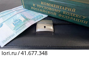 Рублевые купюры лежат на черном деловом портфеле рядом с книгой "Комментарии к трудовому кодексу Российской Федерации" Стоковое фото, фотограф Цибаев Алексей / Фотобанк Лори