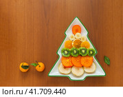 Fresh juicy fruits are sliced in a festive New Year's plate in the shape of a Christmas tree on a wooden table. The concept of proper healthy eating during holiday feasts. Flat lay, closeup, top view, copy space. Стоковое фото, фотограф Виктория Катьянова / Фотобанк Лори