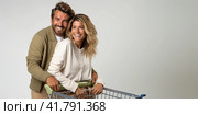 A young couple stands on a gray background with a supermarket cart and smiles. Стоковое AI-изображение, AI-художник Ярославский Максим / Фотобанк Лори