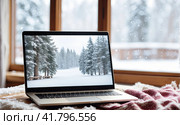 Laptop with a winter landscape screen, indoor on the table with a cozy blanket by the window with snow. Seasonal remote work, internet, shopping, Christmas and New Year. AI generated. Стоковое AI-изображение, AI-художник Ольга Симонова / Фотобанк Лори