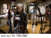 Elderly woman with preteen granddaughter in protective masks examining retro bicycles in museum. Стоковое фото, фотограф Яков Филимонов / Фотобанк Лори