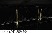 A cable-stayed highway bridge with tall concrete pylons illuminated by architectural lighting at night. General plan of the bridge against the background of the night city lights (2023 год). Редакционное видео, видеограф Даниил Хабаров / Фотобанк Лори